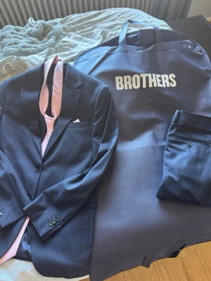 Mörkblå randig kostym Brothers - Snygg mörkblå randig kostym från Brothers med matchande byxor och kavaj. Medföljer en ljusrosa slips från Christensen Sweden och kostymfodral. Klassisk passform och diskreta ränder för en stilren look. Storlek S, endast använd en gång 