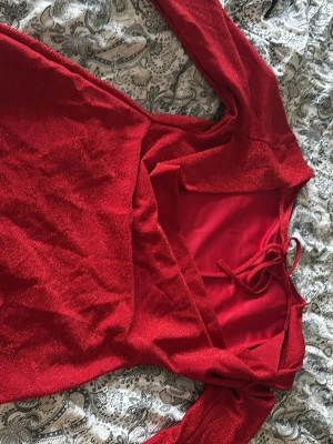 Röd glittrig kortklänning med knyt - Snygg röd kortklänning med glittrigt tyg och långa ärmar. Klänningen har en rund halsringning och knyt i nacken för extra detalj. Perfekt för dig som vill sticka ut med en färgstark och trendig look.