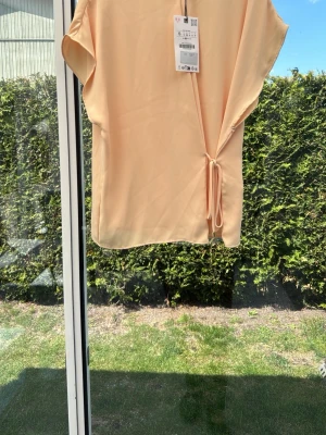 Beige omlottopp med knyt - Säljer en stilren beige omlottopp från Zara med korta ärmar och knyt i sidan. Toppen har en lös passform och är tillverkad i ett mjukt syntetmaterial som känns skönt mot huden. Perfekt för dig som gillar minimalistisk och trendig stil.