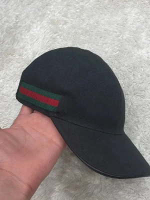 Svart keps från Gucci med röd/grön rand - Snygg svart keps från Gucci med klassisk röd och grön rand på sidan. Kepsen har böjd skärm och justerbart spänne bak. Tillverkad i tyg med diskret GG-mönster i materialet. Perfekt accessoar för en trendig och exklusiv look.(priset kan diskuteras)
