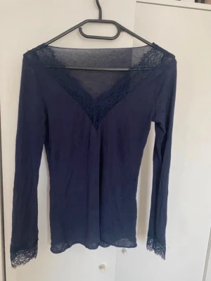 Mörkblå långärmad topp med spets - Snygg mörkblå långärmad topp med v-ringning och spetsdetaljer vid halsringning och ärmslut. Figurnära passform som framhäver siluetten. Perfekt att styla med jeans eller kjol för en trendig look. Storlek M