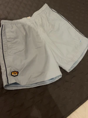 Ljusblå shorts från Nike TN - Snygga ljusblå shorts från Nike TN med svart piping längs sidorna och TN-logga framtill. Shortsen har elastisk midja och är tillverkade i ett lätt syntetmaterial, perfekta för sommaren.