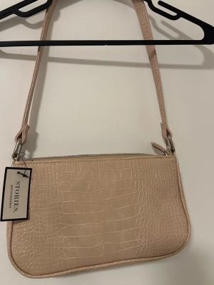 Beige croco handväska från & Other Stories - Stilren beige handväska från & Other Stories med croco-mönstrad yta och silverfärgade detaljer. Väskan har en avlång, mjukt rundad form och justerbar axelrem. Perfekt för dig som gillar trendiga accessoarer i fejkskinn.