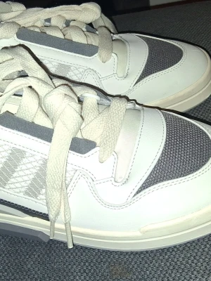 Vita och grå Adidas sneakers - Snygga sneakers från Adidas i vitt och grått med klassiska ränder på sidan. Ovandelen är i en mix av mesh och syntetläder, vilket ger både stil och andningsförmåga. Tjock vit sula och breda vita skosnören för en sportig look.