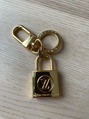 Vuitton guldfärgad nyckelring - Säljer en exklusiv guldfärgad nyckelring från Vuitton med ikoniskt LV-emblem. Nyckelringen har en robust karbinhake och ring, samt ett hänglåsformat hänge i metall. Perfekt accessoar för att piffa till väskan eller nyckelknippan.