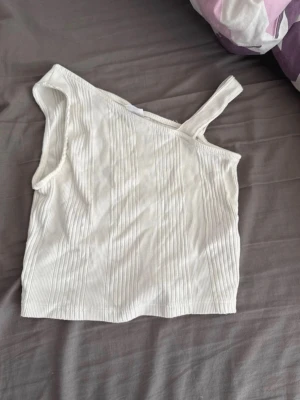 Vit oneshoulder ribbad topp - Snygg vit ribbad topp med oneshoulder-design och ett axelband. Perfekt för dig som vill ha en trendig och stilren look. Materialet är mjukt och stretchigt, vilket gör den bekväm att bära. Passar till många olika outfits och är enkel att matcha. Använd en gång till Taylor swifts konsert. En liten omärkbar fläck