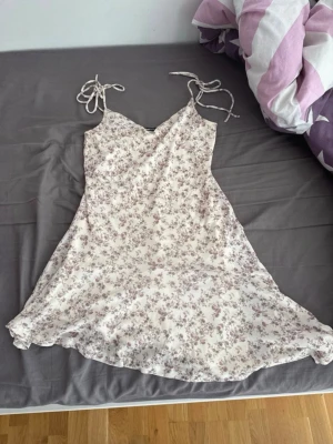 Blommig ljus kortklänning med knyt - Söt ljus kortklänning med blommigt mönster och knytband på axlarna. Klänningen har v-ringad hals och är ärmlös, perfekt för sommarens varma dagar. Materialet känns lätt och luftigt, vilket gör den bekväm att bära.