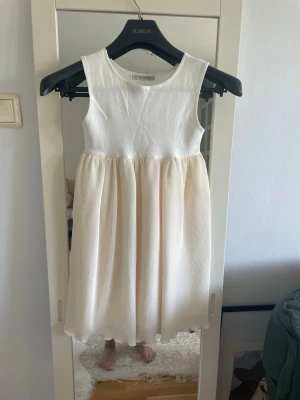 Vit och beige ärmlös klänning - Söt ärmlös klänning med vit överdel i mjuk bomull och plisserad beige kjol i lätt material. Perfekt för sommarens alla tillfällen med sin luftiga och stilrena design. Rund halsringning och midjelinje som markerar övergången mellan materialen.storlek står inte ,men den är 6åring