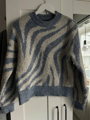 Blåvit zebramönstrad stickad tröja - Säljer en stickad tröja med zebramönster i blått och beige. Tröjan har rund hals, långa ärmar och ribbstickade muddar vid ärmslut och nederkant. Materialet ser mjukt och fluffigt ut, perfekt för kyliga dagar. Använd en del men inte alltför sliten🩷