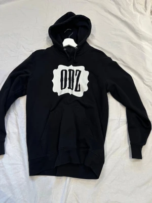 Svart hoodie med ODZ-tryck - Snygg svart hoodie med stort vitt ODZ-tryck på bröstet. Klassisk känguruficka framtill och justerbar huva med snören. Tillverkad i mjukt material som känns skönt mot huden. Perfekt för dig som gillar streetwear och vill sticka ut.