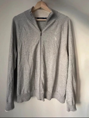 Grå ziptröja från Polo Ralph Lauren - Grå ziptröja från Polo Ralph Lauren med diskret logga broderad på bröstet. Tröjan har hög krage, långa ärmar och ribbade muddar. Tillverkad i mjukt bomullsmaterial och har en klassisk, avslappnad passform.