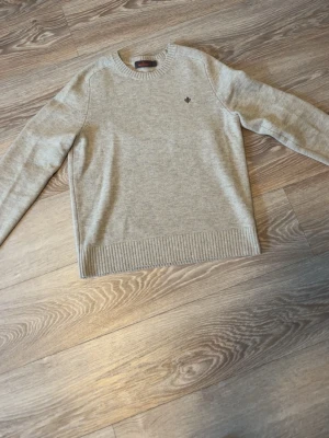 Morris tröja - Snygg beige stickad tröja med rund hals och diskret mörkbrun broderad detalj på bröstet. Tröjan har ribbstickade muddar vid ärmslut och nederkant. Perfekt för lager på lager och enkel att matcha med olika stilar.