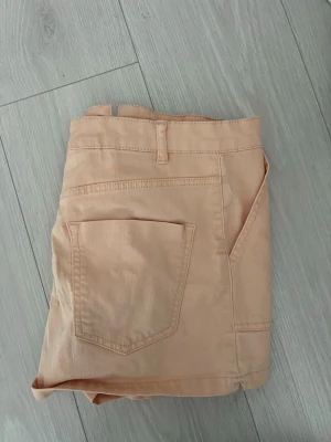 Corall färgade jeansshorts - Säljer ett par corall/aprikos färgade shorts med klassisk design och fickor både fram och bak. Shortsens passform är normal och de har hällor för bälte samt knapp och dragkedja framtill. Perfekta för varma dagar och enkla att matcha med olika toppar. Jag brukar vika ner kanten så dom blir lågmidjade!
