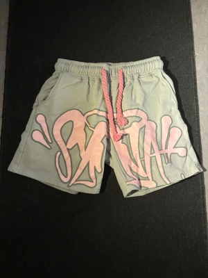 Syna world shorts - Säljer ett par gråa shorts med stort rosa tryck framtill. Shortsen har resår i midjan och rosa snörning för justerbar passform.  