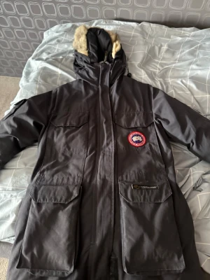 Svart parkas från Canada Goose - Säljer en svart parkasjacka från Canada Goose med luva och fuskpälsdetalj. Jackan har flera stora fickor framtill, dragkedja och tryckknappar. Klassisk Canada Goose-logga på bröstet och patch på fickan. Perfekt för kalla dagar.