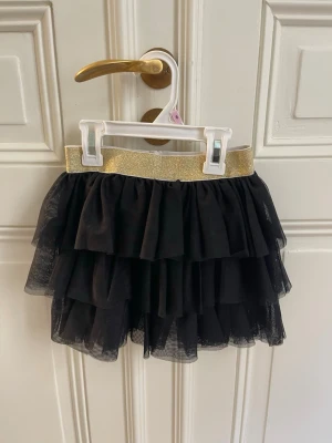 Svart volangkjol med guldresår Palomino - Supersöt svart kortkjol från Palomino med flera lager volanger och bred glittrig guldresår i midjan. Kjolen är gjord i ett lätt, skirt material som ger ett fluffigt intryck och passar perfekt för att sticka ut lite extra.