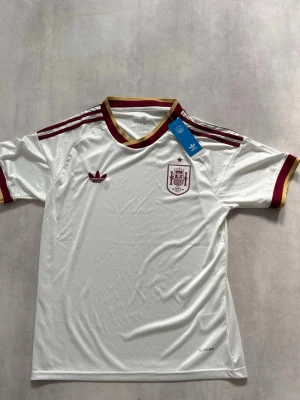 Vit Spanien tröja VM 2026 - Vit fotbollströja från Adidas med Spaniens landslagsmärke på bröstet. Tröjan har vinröda och guldiga detaljer på krage, ärmslut och axlar. Materialet är lätt och andas, perfekt för sport och träning. Klassisk Adidas-logo på framsidan.