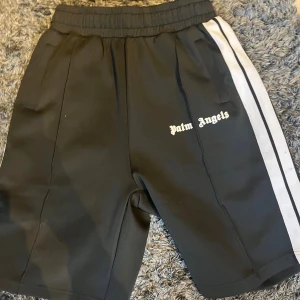 Palm Angels svarta shorts med vita ränder - Svarta shorts från Palm Angels med vita sidoremsor och logotyp tryckt på vänster ben. Modellen har elastisk midja och två fickor fram. Tillverkade i 100% polyester för en sportig vibe och skön passform. Perfekta för dig som gillar streetwear och snygga detaljer. Jag vet inte om de är äkte eller om de är kopior. Vet ej om de är riktiga
