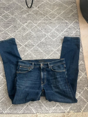 Mörkblå jeans från Nudie Jeans - Säljer ett par klassiska mörkblå jeans från Nudie Jeans med raka ben och fem fickor. Jeansen har kontrastsömmar och knappgylf. Perfekta för dig som gillar en tidlös och clean look. Materialet är slitstark denim i bomull. Har haft ett hål i skrevet men har blivit igensytt av skreddare (bild4)