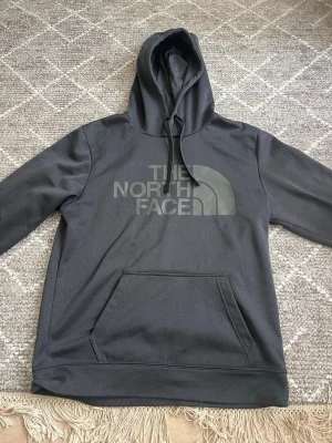 Svart hoodie från The North Face - Snygg svart hoodie från The North Face med stor logga framtill. Klassisk modell med huva och känguruficka. Tillverkad i mjukt och bekvämt material, perfekt för chill eller streetwear. Dragsko i huvan för justerbar passform.