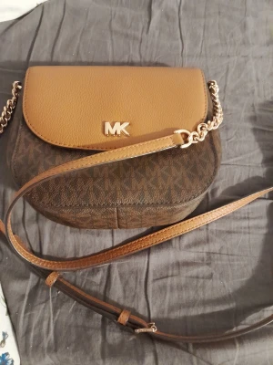 Brun axelväska från Michael Kors - Snygg axelväska från Michael Kors i brunt konstläder med logomönster och guldfärgade detaljer. Väskan har en justerbar axelrem med kedjedetaljer och ett lock med MK-logga framtill. Perfekt storlek för det viktigaste och stilren design.