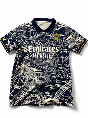 Real Madrid Adidas drakmönstrad tröja - Snygg Real Madrid-tröja från Adidas med mörkblå bas och unikt drakmönster i vitt. Tröjan har klassisk krage, klubbmärke på bröstet och gula detaljer på ärmsluten. Perfekt för dig som vill sticka ut på planen eller läktaren.