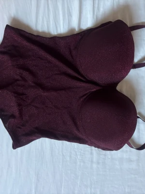 Vinröd glittrig body med kupor - Snygg vinröd body med glittrigt tyg och vadderade kupor. Justerbara axelband och figurnära passform. Perfekt för dig som vill ha något extra under kläderna. Materialet är stretchigt och ger en lyxig känsla.