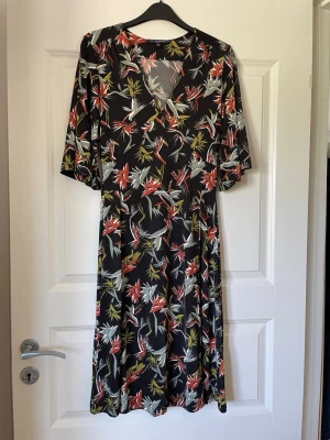 Blommig knälång klänning Ilse Jacobsen - Svart klänning från Ilse Jacobsen med blommigt mönster i rött, vitt och gult. Klänningen har v-ringning och halvlånga ärmar. Perfekt för dig som gillar färgstarka prints och en avslappnad men stilren look. Skön att bära!
