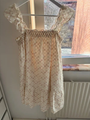 Sommarklänning - Söt kortklänning i ljusbeige med blommigt broderat mönster och volangdetaljer. Klänningen har breda axelband med volanger och smock upptill för en bekväm passform. Perfekt för sommarens alla tillfällen!
