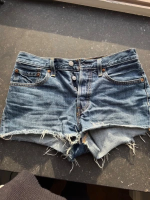 Levi's jeansshorts med fransig kant - Snygga blå jeansshorts från Levi's med fransig, rå kant och klassisk femficksdesign. Shortsen har hög midja, 3 knappar, avslappnad look. Perfekta för sommardagar, strlk 36/38 passar inte längre
