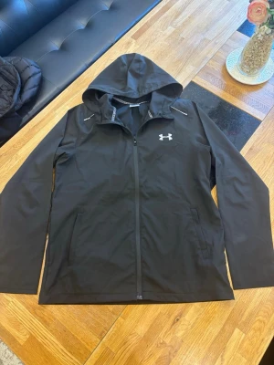 Under Armor Windbreaker  - Snygg svart vindjacka från Under Armour med huva och dragkedja framtill. Jackan har två sidofickor, vita detaljer vid axlarna och logga på bröstet. Tillverkad i lätt och funktionellt material, perfekt för blåsiga dagar.