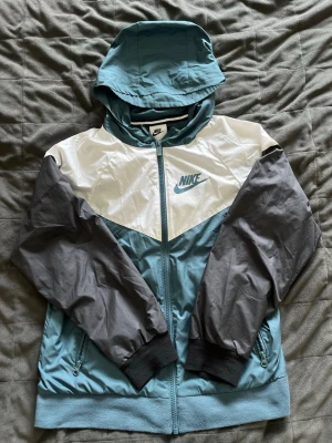 Nike vindjacka med huva blå/vit/grå - Snygg vindjacka från Nike i blått, vitt och grått. Jackan har huva, dragkedja framtill, två sidofickor med dragkedja och muddar vid ärmsluten. Nike-logga på bröstet. Perfekt för blåsiga dagar och sportig stil.