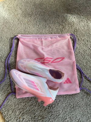 Rosa Adidas X lamine yamal f50 - Säljer ett par Adidas X Speedflow fotbollsskor i en snygg rosa och vit färgkombination. Skorna har en strömlinjeformad design utan snörning och är tillverkade i lätt syntetmaterial för maximal fart på planen. Medföljer gör en matchande rosa meshpåse.