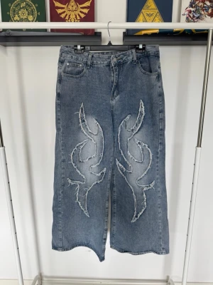 Blå wide leg jeans - Säljer ett par blå wide leg jeans med råa flames-detaljer framtill. Jeansen har hög midja, fem fickor och en avslappnad passform. Materialet är klassisk denim i bomull och färgen är tvättad ljusblå. 
