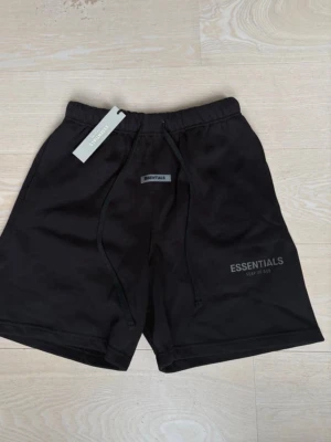 Svarta shorts från Essentials - Säljer ett par svarta shorts från Essentials med snörning i midjan och diskret logga på benet. Shortsen har en avslappnad passform och är perfekta för en chill stil. Materialet känns mjukt och bekvämt, troligen bomull eller bomullsblandning.