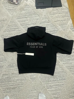 Svart hoodie Essentials Fear of God - Snygg svart hoodie från Fear of God Essentials med stor logga på ryggen och diskret patch framtill. Klassisk känguruficka och rejäl huva. Tillverkad i mjuk bomullsblandning för maximal komfort. Perfekt för en clean och trendig streetwear-look.