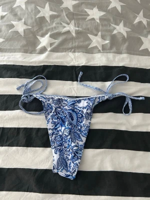 Blåvit bikiniunderdel med knyt - Säljer en snygg bikiniunderdel i blått och vitt med blommigt mönster. Modellen har smala knytband i sidorna för justerbar passform och är perfekt för strandhäng. Materialet känns mjukt och följsamt. Helt oanvänd 