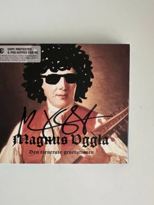 Signerad Magnus Uggla CD - ”Den tatuerade generationen” - Signerad CD av Magnus Uggla : ”Den tatuerade generationen". Den innehåller 11 spår, inklusive "Magnuspassionen" och "Värsta grymma tjejen". Skivan kommer i ett utvikbart fodral med bilder på en pudel med solglasögon och peruk. Mycket gott skick, som ny!
