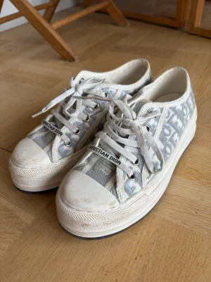 Christian Dior vita låga sneakers - Snygga vita låga sneakers från Christian Dior med klassiskt monogrammönster i grått och vitt. Skorna har vita snören, robust sula och detaljer med logotyp på plösen. Tillverkade i canvasmaterial med gummisula. Perfekta för en trendig och avslappnad stil. Orginalförpackning finns kvar:) syntesen borde enkelt gå bort 