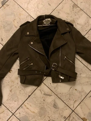 Olivgrön skinnjacka med bälte - Snygg olivgrön skinnjacka i bikerstil med silvriga dragkedjor, axelklaffar och bälte i midjan i storlek M men passar om man har storlek S. Jackan har flera fickor med dragkedja och en klassisk krage. Perfekt för dig som vill ha en cool och trendig look. Har använts några gånger och har en knapp som saknas, men annars jättebra skick! 