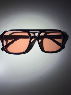 Svarta solglasögon med rosa glas - Snygga solglasögon med svart, kantig båge och rosa tonade glas. Modellen har en retroinspirerad form med bred brygga och raka skalmar. Perfekta för dig som vill sticka ut med en cool accessoar till sommaren.