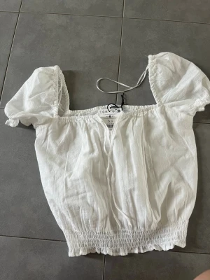 Vit puffärm blus från Bershka - Supersöt vit blus från Bershka med puffiga korta ärmar och smockad nederkant. Toppen har en luftig och lätt känsla, perfekt för varma dagar. OBS! kommer ej sänka priset utan säljer den för vad jag köpte den för pga att den är oanvänd