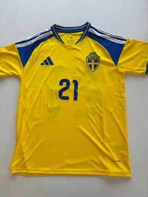 Sverige matchtröja KULUSEVSKI #21 - Gul och blå Sverige matchtröja från Adidas med nummer 21 och namnet KULUSEVSKI på ryggen. Tröjan har blå detaljer på axlar och sidor, samt det svenska landslagets emblem på bröstet. Tillverkad i lätt och ventilerande material, perfekt för fotboll.
