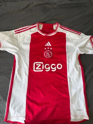 Ajax fotbollströja Adidas röd/vit - Ajax hemmatröja från Adidas i klassiskt röd och vit design. Tröjan har Ziggo-logga på bröstet, klubbmärke och tre stjärnor ovanför. Korta ärmar, rund hals och tre röda ränder på axlarna. Tillverkad i lätt och ventilerande material, perfekt för fotboll.