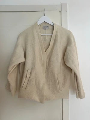 Beige cardiganjacka från ÀVORA - Stilren beige cardiganjacka från ÀVORA med rak passform och djup V-ringning. Jackan har två fickor framtill och är tillverkad i ett mjukt, filtliknande material som ger en avslappnad men ändå snygg look. Perfekt att slänga över en t-shirt eller hoodie.