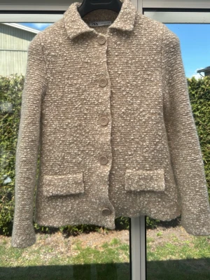 Beige bouclékofta från Zara - Säljer en beige bouclékofta från Zara med stora knappar framtill och två fickor. Koftan har en klassisk krage och är tillverkad i ett mjukt, strukturerat material som ger en trendig look. Perfekt för lager-på-lager-stil.