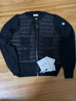 Svart cardiganjacka från Moncler - Snygg svart cardiganjacka från Moncler med quiltad front och stickade ärmar. Dragkedja framtill och ribbade muddar. Ikonisk Moncler-logga på ärmen och två fickor fram. Perfekt för lager på lager och streetstyle. Kvitto,tags och påse medföljer😊