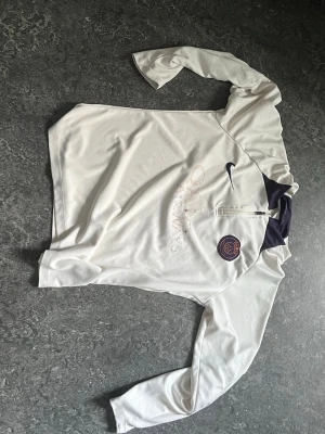 Vit Nike PSG långärmad träningströja - Säljer en vit långärmad träningströja från Nike med PSG-logga på bröstet och mörkblå detaljer vid kragen. Tröjan har halv dragkedja framtill och är tillverkad i ett lätt, sportigt material som passar perfekt till träning eller uppvärmning.
