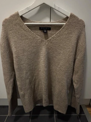 Beige stickad tröja med v-ringning - Mysig beige stickad tröja från AMISU med v-ringad hals och långa ärmar. Tröjan har en mjuk, fluffig struktur och ribbstickade muddar vid ärmslut och nederkant. Perfekt för dig som gillar en enkel och stilren look.