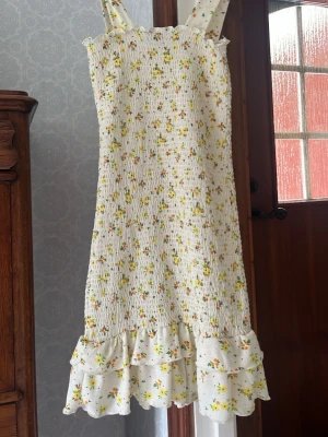 Blommig sommarklänning med volang - Söt vit klänning med små gula och orange blommor, breda axelband och volangkant nertill. Smockad överdel ger en bekväm passform. Perfekt för varma dagar och har en romantisk känsla. 